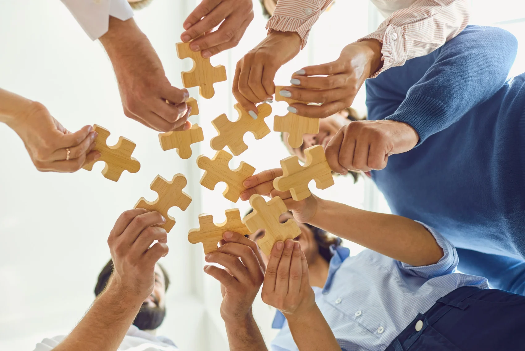 Several hands each holding a wooden puzzle piece towards a centre, representing how separate experiences may form a recognisable pattern